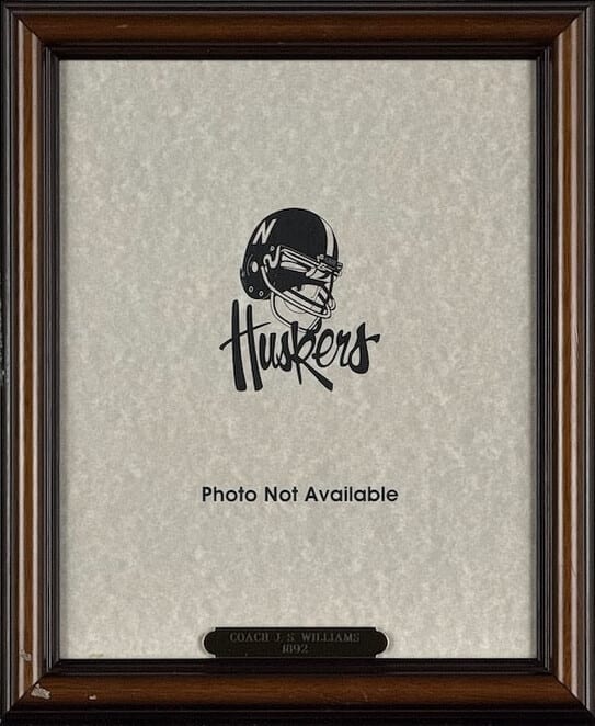 A wood frame holding a card with the Huskers logo and the words Photo Not Available, above a brass plate for Coach J.S. Williams, 1892.
