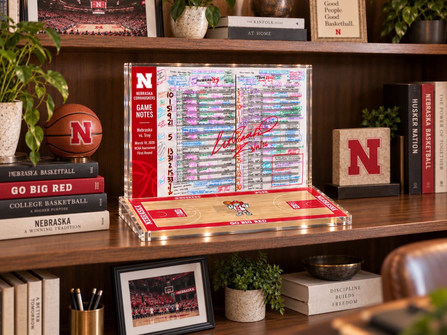 The display standing on a wooden bookshelf beside Nebraska basketball books, a leather basketball with the Nebraska N, framed photographs of a packed arena and a small potted plant. The Troy board faces out, signed in red, on its court base.