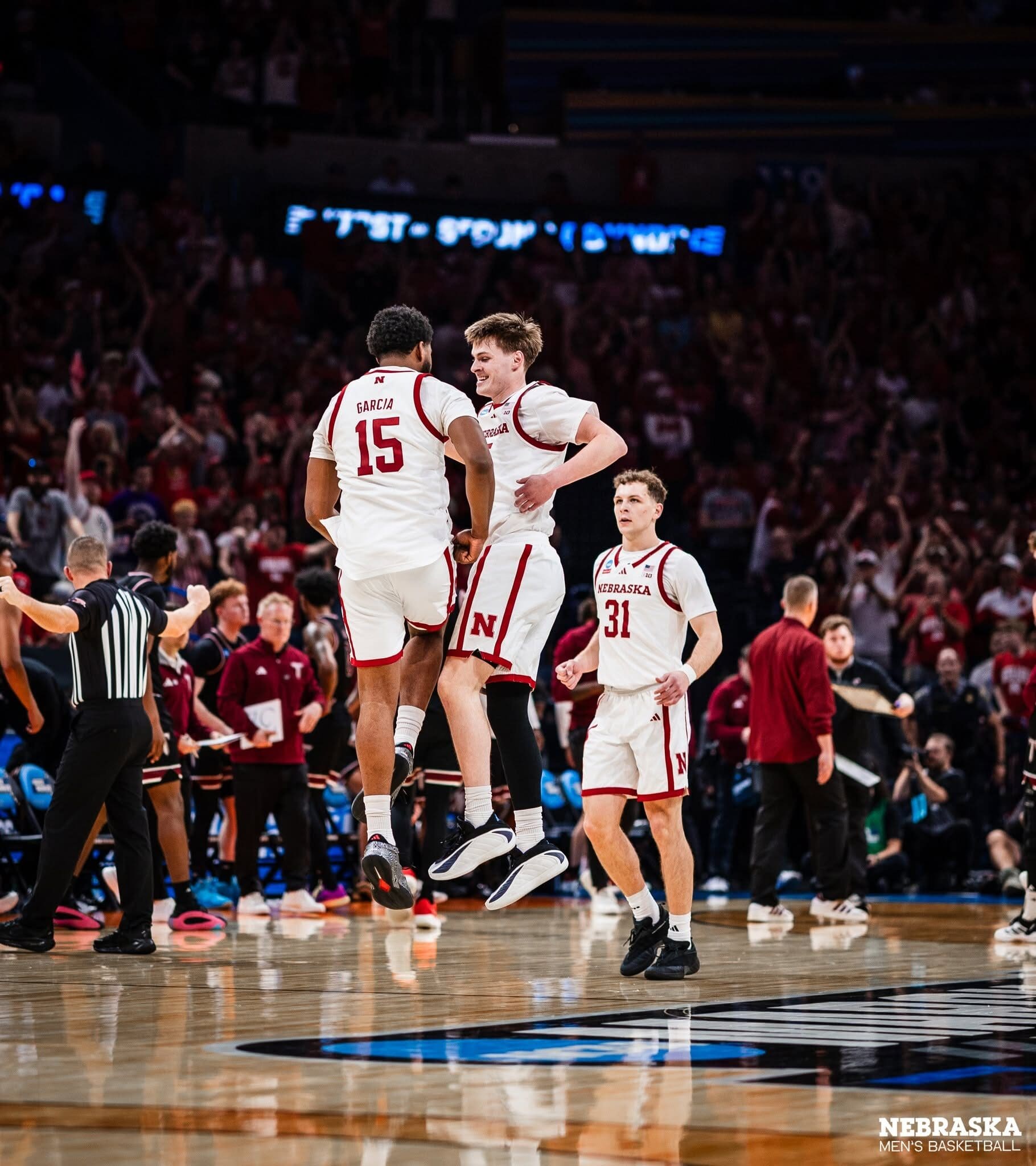 Two Nebraska players leaping into each other mid-court in celebration, number 15 and a teammate, with number 31 running in behind them and a referee at the edge of the frame. The stand behind is a wall of red, every seat standing.