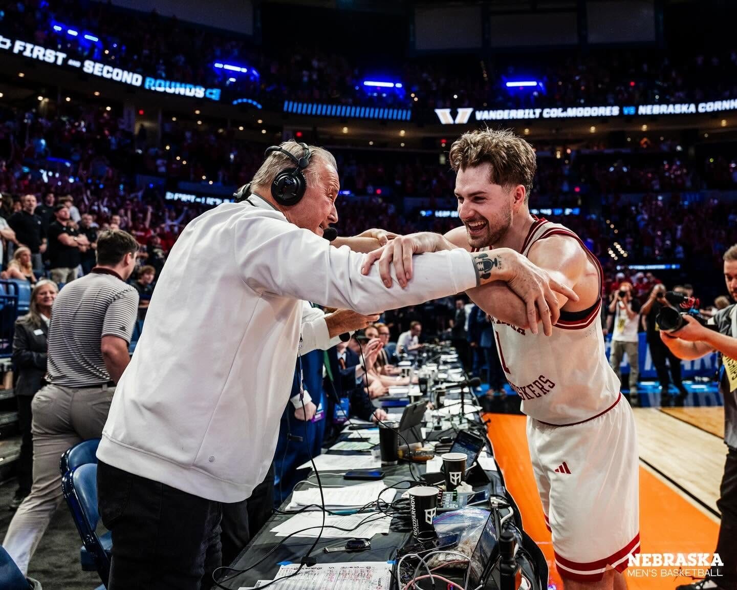 Kent Pavelka at the courtside broadcast table in a white quarter-zip and headset, reaching across the table to clasp arms with a grinning Nebraska player in a white Huskers jersey. Behind them the arena screen reads “Vanderbilt Commodores vs Nebraska Cornhuskers”, under “First and Second Rounds” signage, with a packed house standing and photographers crowding in at the right.